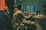 A person is seated at a desk with a computer, working on music software. The room is equipped with acoustic panels, headphones, a keyboard, and speakers. The setting suggests a studio environment.