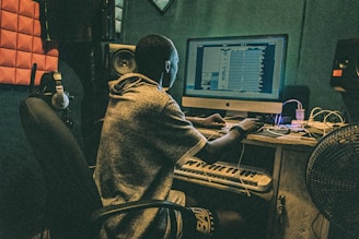 A person is seated at a desk with a computer, working on music software. The room is equipped with acoustic panels, headphones, a keyboard, and speakers. The setting suggests a studio environment.