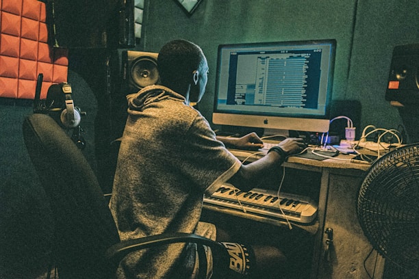 A person is seated at a desk with a computer, working on music software. The room is equipped with acoustic panels, headphones, a keyboard, and speakers. The setting suggests a studio environment.