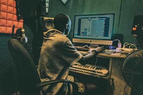 A person is seated at a desk with a computer, working on music software. The room is equipped with acoustic panels, headphones, a keyboard, and speakers. The setting suggests a studio environment.