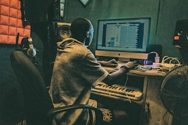 A person is seated at a desk with a computer, working on music software. The room is equipped with acoustic panels, headphones, a keyboard, and speakers. The setting suggests a studio environment.