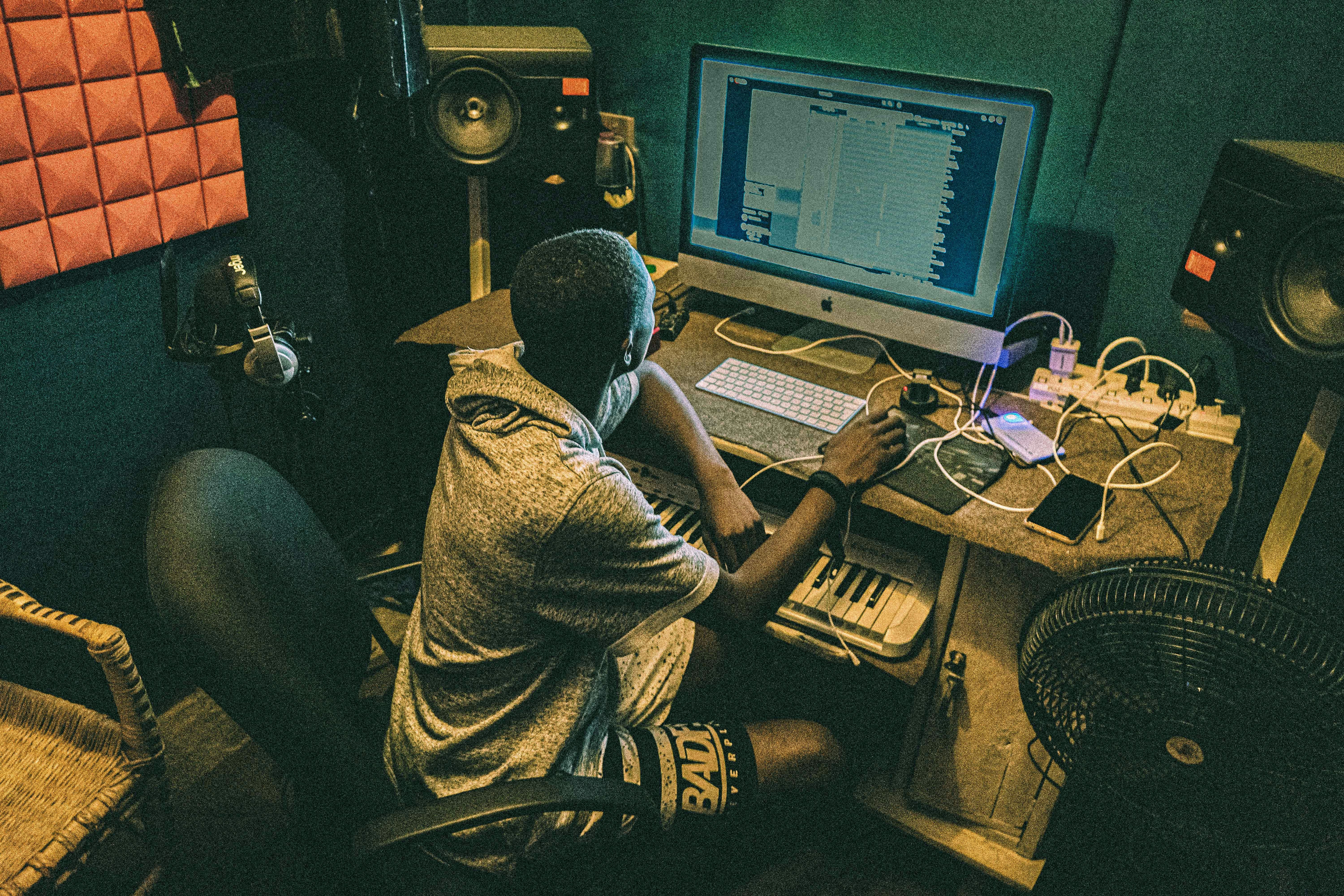 A person is seated at a desk in a music production studio, working on a computer. The setup includes speakers, a pop filter microphone, and an interface with a keyboard. The lighting is dim, creating a focused atmosphere.