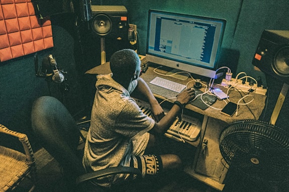 A person is seated at a desk in a music production studio, working on a computer. The setup includes speakers, a pop filter microphone, and an interface with a keyboard. The lighting is dim, creating a focused atmosphere.