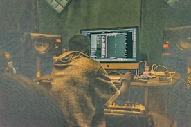 A person is sitting at a desk in front of a large computer monitor, working on music production software. The room is filled with audio equipment, including large speakers on either side of the monitor and various cables and electronic devices on the desk.