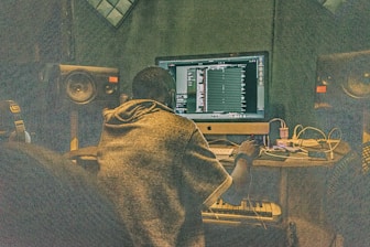 A person is sitting at a desk in front of a large computer monitor, working on music production software. The room is filled with audio equipment, including large speakers on either side of the monitor and various cables and electronic devices on the desk.