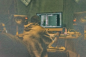 A person is sitting at a desk in front of a large computer monitor, working on music production software. The room is filled with audio equipment, including large speakers on either side of the monitor and various cables and electronic devices on the desk.