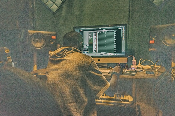 A person is sitting at a desk in front of a large computer monitor, working on music production software. The room is filled with audio equipment, including large speakers on either side of the monitor and various cables and electronic devices on the desk.