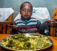 A young child sits at a table with a large metal plate of food. The child is wearing a striped sweater and holding a spoon, looking directly at the camera. The plate contains a green vegetable dish mixed with a starchy food. The setting appears to be indoors with wooden chairs and colorful fabrics in the background.