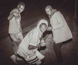 Three young men are posing on a street at night. One is standing with a playful hand gesture, another in the middle is wearing a hooded sweatshirt while slightly crouching, and the third has a jacket and beanie, appearing cheerful. The scene is captured in a monochrome or sepia tone, adding a vintage feel.