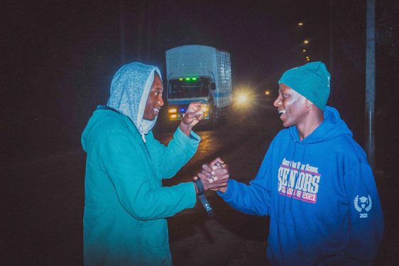 Two friends shaking hands in front of Roya Auto dealership with a row of clean, shiny used cars.