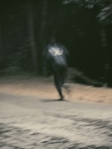 A blurred image of a person running along a dimly lit road surrounded by trees. The motion blur adds a sense of speed and mystery to the scene, enhanced by the low lighting conditions indicating it might be nighttime or dusk.