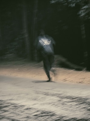 A blurred image of a person running along a dimly lit road surrounded by trees. The motion blur adds a sense of speed and mystery to the scene, enhanced by the low lighting conditions indicating it might be nighttime or dusk.