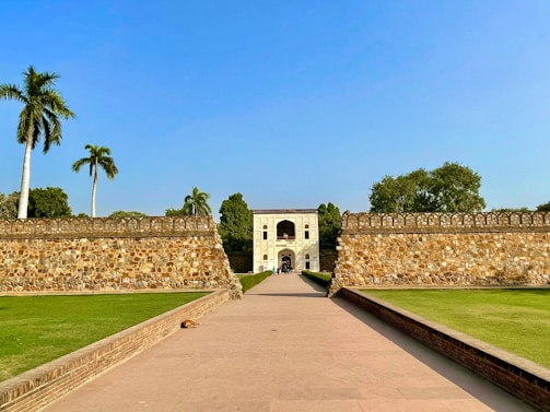 A historical stone gateway stands at the center, framed by two ancient stone walls. The pathway leading to the gateway is lined with neatly trimmed green grass on either side. Tall palm trees rise above the walls, and a clear blue sky stretches above.