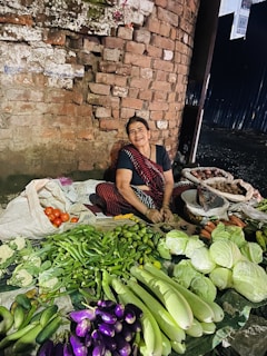 A woman, dressed in traditional attire, is sitting on the ground in front of a brick wall and selling a variety of fresh vegetables, including cabbages, eggplants, cucumbers, tomatoes, and more. The vegetables are neatly arranged on cloth or plastic sheets.