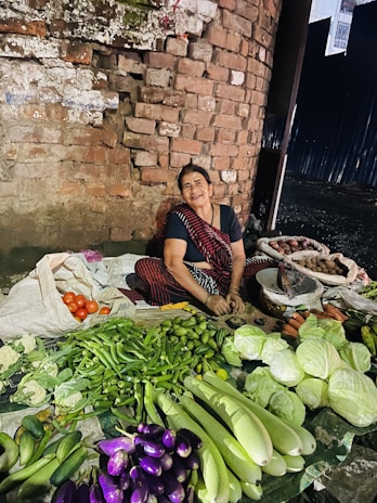 A woman, dressed in traditional attire, is sitting on the ground in front of a brick wall and selling a variety of fresh vegetables, including cabbages, eggplants, cucumbers, tomatoes, and more. The vegetables are neatly arranged on cloth or plastic sheets.