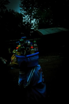 A person carrying a basket on their head, filled with various bottles and packaged goods, against a dark, outdoor background with trees and a building visible.