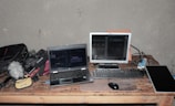 A workspace featuring a laptop, a monitor, a keyboard, and a computer mouse on a wooden table. Various electronic equipment including cables, a microphone with a furry windscreen, and audio gear are scattered around. The setting suggests a focus on audio or video production.