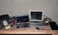 A workspace featuring a laptop, a monitor, a keyboard, and a computer mouse on a wooden table. Various electronic equipment including cables, a microphone with a furry windscreen, and audio gear are scattered around. The setting suggests a focus on audio or video production.