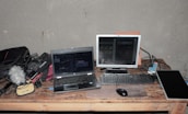 A workspace featuring a laptop, a monitor, a keyboard, and a computer mouse on a wooden table. Various electronic equipment including cables, a microphone with a furry windscreen, and audio gear are scattered around. The setting suggests a focus on audio or video production.