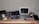 A workspace featuring a laptop, a monitor, a keyboard, and a computer mouse on a wooden table. Various electronic equipment including cables, a microphone with a furry windscreen, and audio gear are scattered around. The setting suggests a focus on audio or video production.