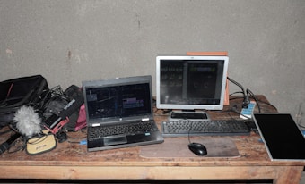 A workspace featuring a laptop, a monitor, a keyboard, and a computer mouse on a wooden table. Various electronic equipment including cables, a microphone with a furry windscreen, and audio gear are scattered around. The setting suggests a focus on audio or video production.