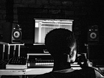 A person is sitting in front of a music production setup, with a computer screen displaying a digital audio workstation interface. Surrounding the individual are speakers and audio equipment, indicative of a studio environment.