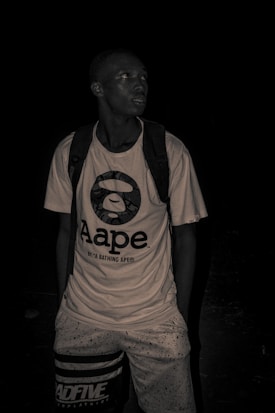 A person is wearing a graphic t-shirt with the Aape logo and a backpack. The image is dark and has a moody, monochrome appearance.