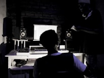 A dimly lit recording studio with two people present. One person is seated at a desk with a computer screen displaying audio editing software. Studio speakers and a MIDI keyboard are positioned on the desk. The second person stands beside the desk, wearing headphones around the neck.