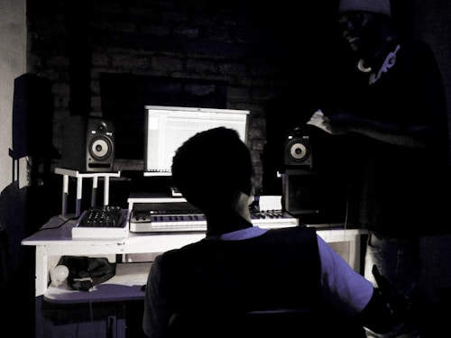 A dimly lit recording studio with two people present. One person is seated at a desk with a computer screen displaying audio editing software. Studio speakers and a MIDI keyboard are positioned on the desk. The second person stands beside the desk, wearing headphones around the neck.