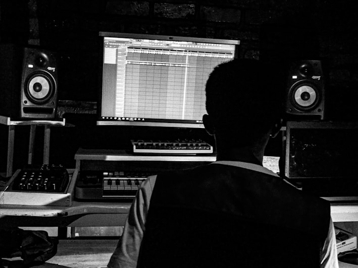 A person is seated at a music production setup, featuring a computer screen displaying music editing software. The setup includes speakers on either side of the desk and various music production equipment.