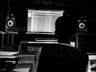 A person is seated at a music production setup, featuring a computer screen displaying music editing software. The setup includes speakers on either side of the desk and various music production equipment.