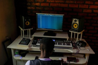 A person is seated at a music production workstation featuring a computer monitor displaying a digital audio workstation (DAW) interface. The setup includes a keyboard, two studio monitors with yellow speaker cones, a mixer, headphones, and various electronic equipment on a desk. The background features an exposed brick wall.