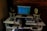 A person is seated at a music production workstation featuring a computer monitor displaying a digital audio workstation (DAW) interface. The setup includes a keyboard, two studio monitors with yellow speaker cones, a mixer, headphones, and various electronic equipment on a desk. The background features an exposed brick wall.