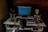 A person is seated at a music production workstation featuring a computer monitor displaying a digital audio workstation (DAW) interface. The setup includes a keyboard, two studio monitors with yellow speaker cones, a mixer, headphones, and various electronic equipment on a desk. The background features an exposed brick wall.