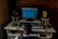 A person is seated at a music production workstation featuring a computer monitor displaying a digital audio workstation (DAW) interface. The setup includes a keyboard, two studio monitors with yellow speaker cones, a mixer, headphones, and various electronic equipment on a desk. The background features an exposed brick wall.