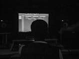 A person sits in a dark room facing a computer screen displaying music production software. The setup includes speakers and other audio equipment, creating a studio-like environment.