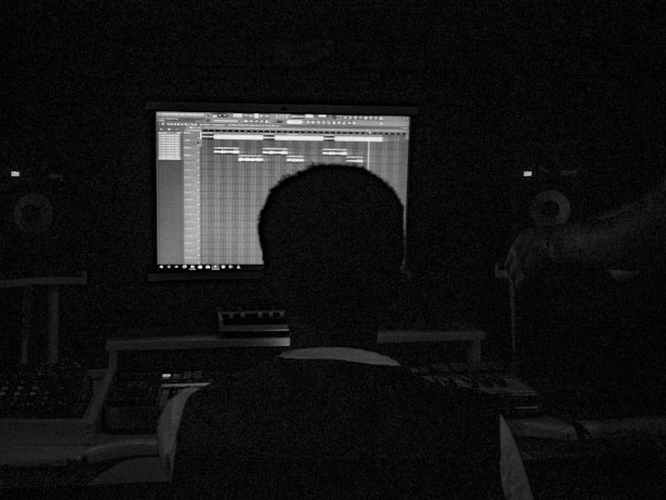 A person sits in a dark room facing a computer screen displaying music production software. The setup includes speakers and other audio equipment, creating a studio-like environment.