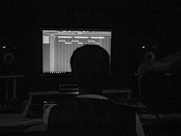 A person sits in a dark room facing a computer screen displaying music production software. The setup includes speakers and other audio equipment, creating a studio-like environment.