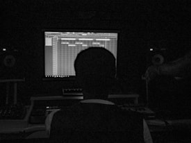 A person sits in a dark room facing a computer screen displaying music production software. The setup includes speakers and other audio equipment, creating a studio-like environment.