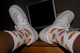 Feet are propped up on a wooden surface, wearing white sneakers and socks with a colorful pattern of red and yellow peppers. In the background, there is a computer monitor with a blank, dark screen.