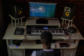 A person is seated at a music production workstation. The setup includes a keyboard, two speakers, computer monitor displaying a music production software, headphone placed on the desk and various electronic equipment. The room has a dim, cozy lighting.