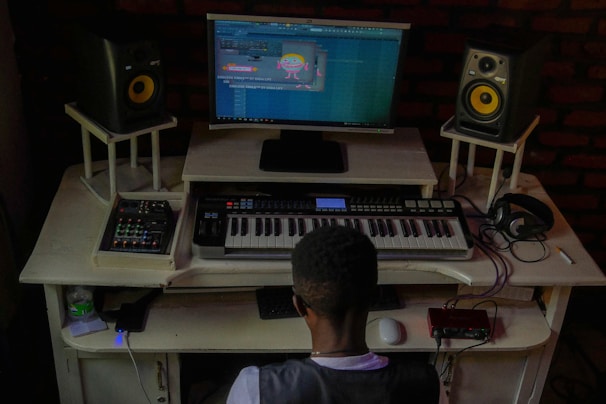 A person is seated at a music production workstation. The setup includes a keyboard, two speakers, computer monitor displaying a music production software, headphone placed on the desk and various electronic equipment. The room has a dim, cozy lighting.
