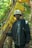 A person in a black jacket and a light-colored bucket hat stands in a forested area. They are holding onto a large, yellowing banana leaf, surrounded by lush green foliage and trees.