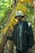 A person in a black jacket and a light-colored bucket hat stands in a forested area. They are holding onto a large, yellowing banana leaf, surrounded by lush green foliage and trees.