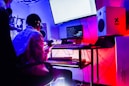 A music studio setup with a person sitting in front of a keyboard and computer screens. The room is dimly lit with neon lights, creating a vibrant atmosphere. Audio equipment, including speakers and a microphone, is visible. The person appears to be engaged in music production, holding a microphone.