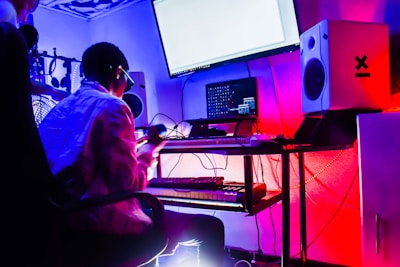 A music studio setup with a person sitting in front of a keyboard and computer screens. The room is dimly lit with neon lights, creating a vibrant atmosphere. Audio equipment, including speakers and a microphone, is visible. The person appears to be engaged in music production, holding a microphone.