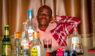 A person is sitting behind a table filled with various alcohol bottles. The individual is wearing a pink hoodie under a red plaid jacket and has a relaxed pose with hands behind the head. The background features brown curtains.