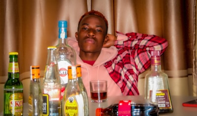A person is sitting behind a table filled with various alcohol bottles. The individual is wearing a pink hoodie under a red plaid jacket and has a relaxed pose with hands behind the head. The background features brown curtains.