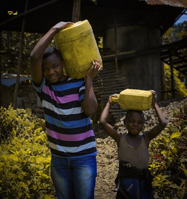 A man and a child are carrying yellow jerrycans on their heads, walking along a path in a rural setting. The man is wearing a striped T-shirt and jeans, while the child has a small smile. The background features a rustic, corrugated building and some foliage.