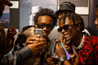 A group of young men are gathered closely together, all wearing stylish attire and sunglasses. They appear to be looking at something on a smartphone with expressions of excitement and amusement. The environment seems casual and social.